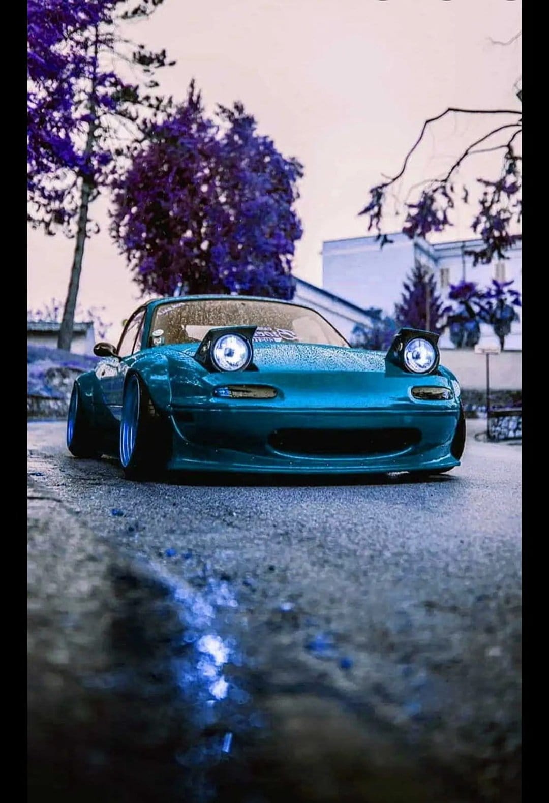 Cyan Mazda Miata with glowing pop-up headlights parked on a wet street with purple trees in the background.