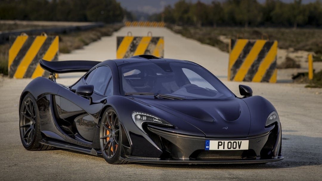 Black McLaren P1 hypercar with orange brake calipers on a grey track between yellow and black barriers.