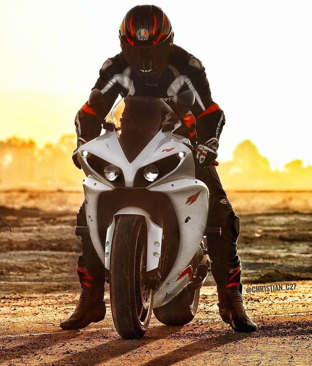 Motorcycle racer in full gear on a white sportbike against a glowing orange and yellow sunset sky backdrop.