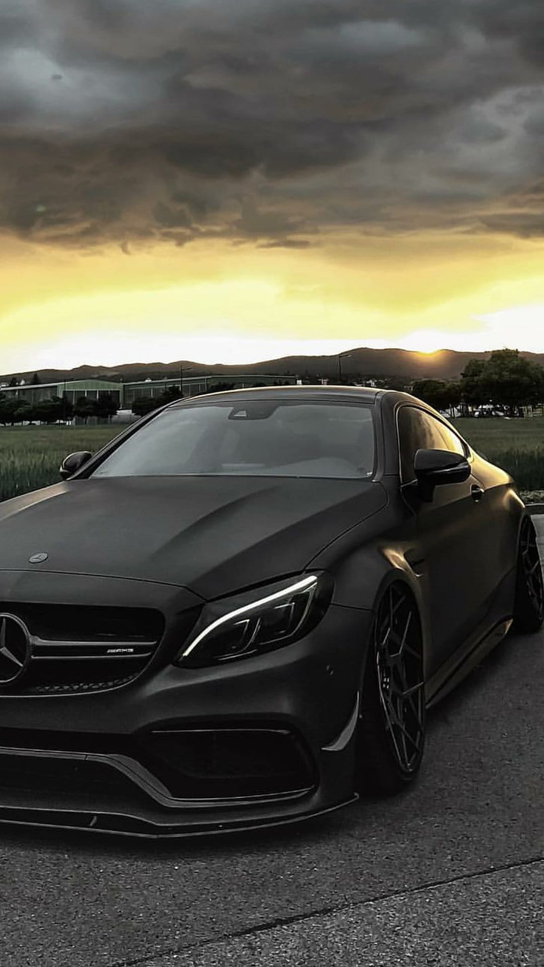 Mercedes-AMG C63 Coupe in matte black parked on asphalt with a dramatic orange sunset sky and hills behind it.