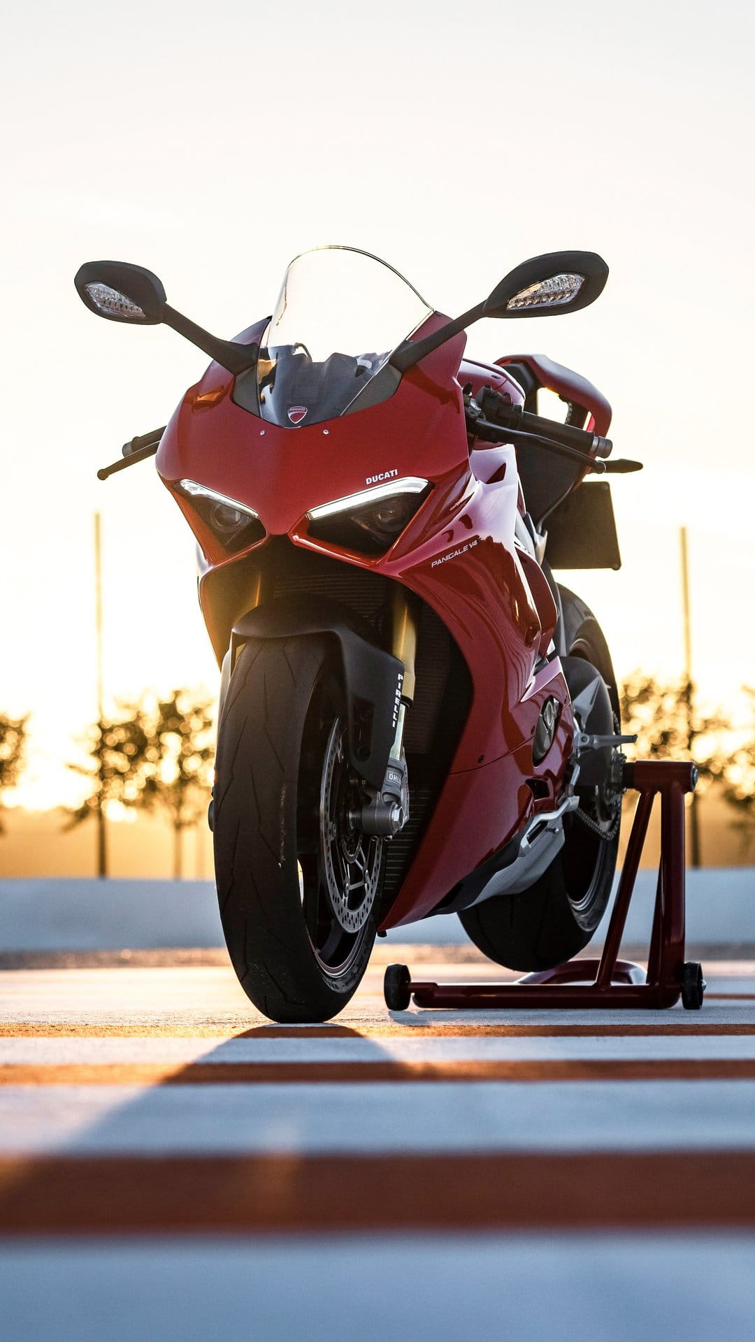 Red Ducati sportbike on a rear stand against a sunset sky with blurred trees and orange striped pavement.