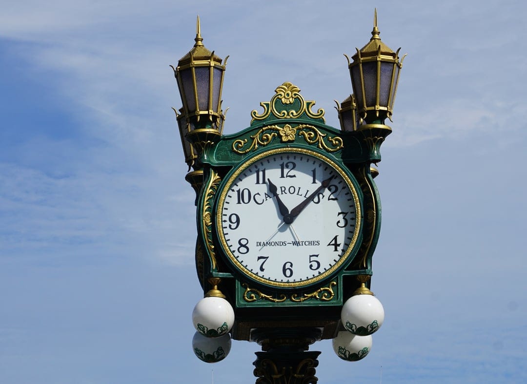 Victorian green and gold street clock featuring Roman numerals and flanking lampposts against a clear blue sky.