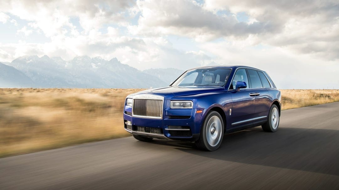 Blue Rolls-Royce Cullinan driving on an open road with majestic mountains and golden fields under a cloudy sky.