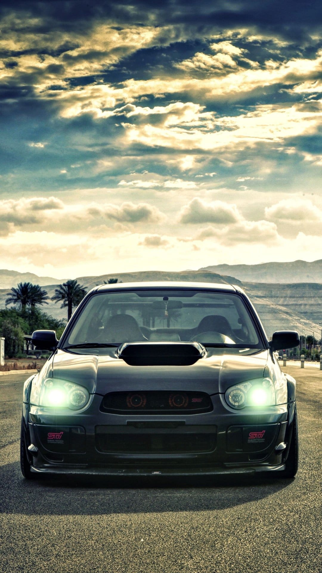 Dark Subaru WRX STI with glowing headlights on a desert road under a dramatic cloudy sky at sunset.