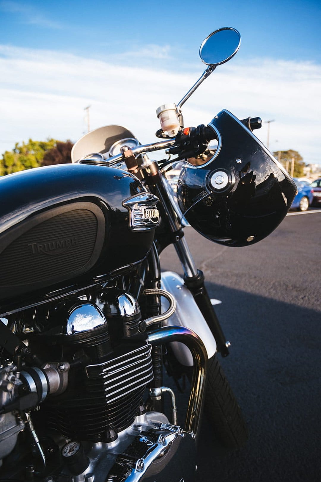 Black Triumph motorcycle with a helmet on the handlebars against a bright blue sky and asphalt road surface.