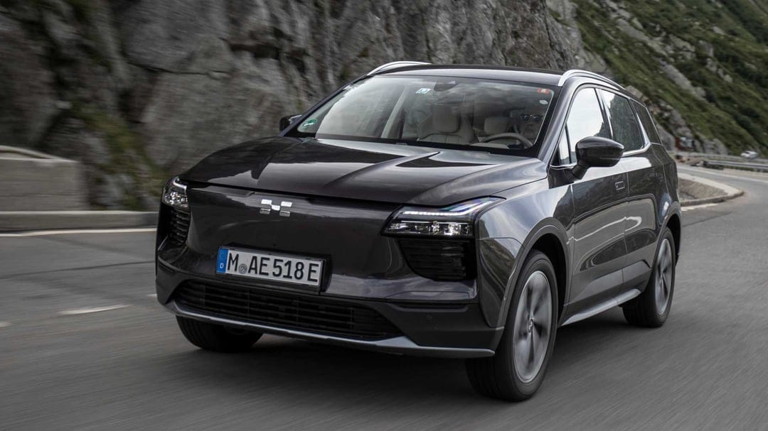 Dark grey electric SUV driving on a winding mountain road with rocky cliffs and motion blurred asphalt.