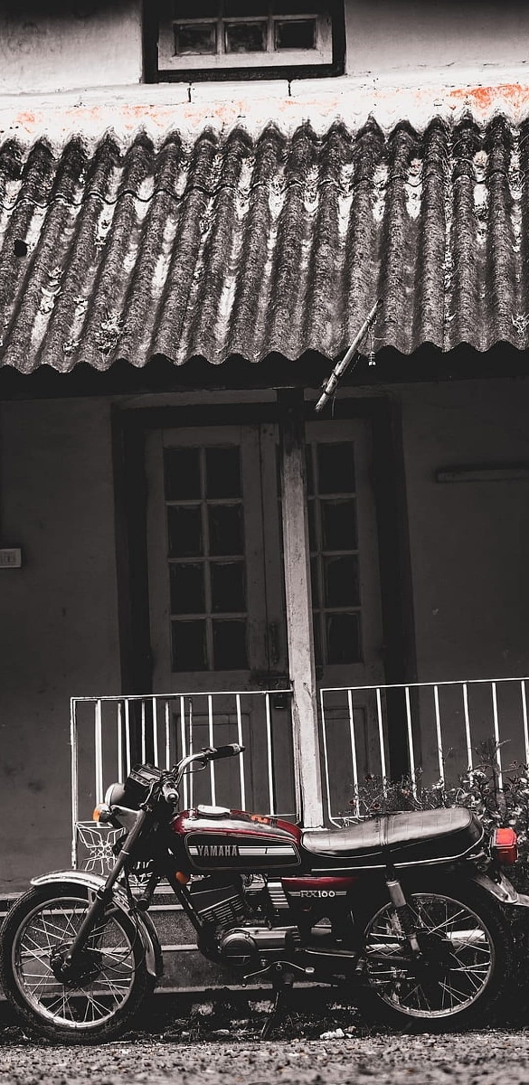 Yamaha RX100 motorcycle parked beside a weathered house with a corrugated metal roof under soft daylight.