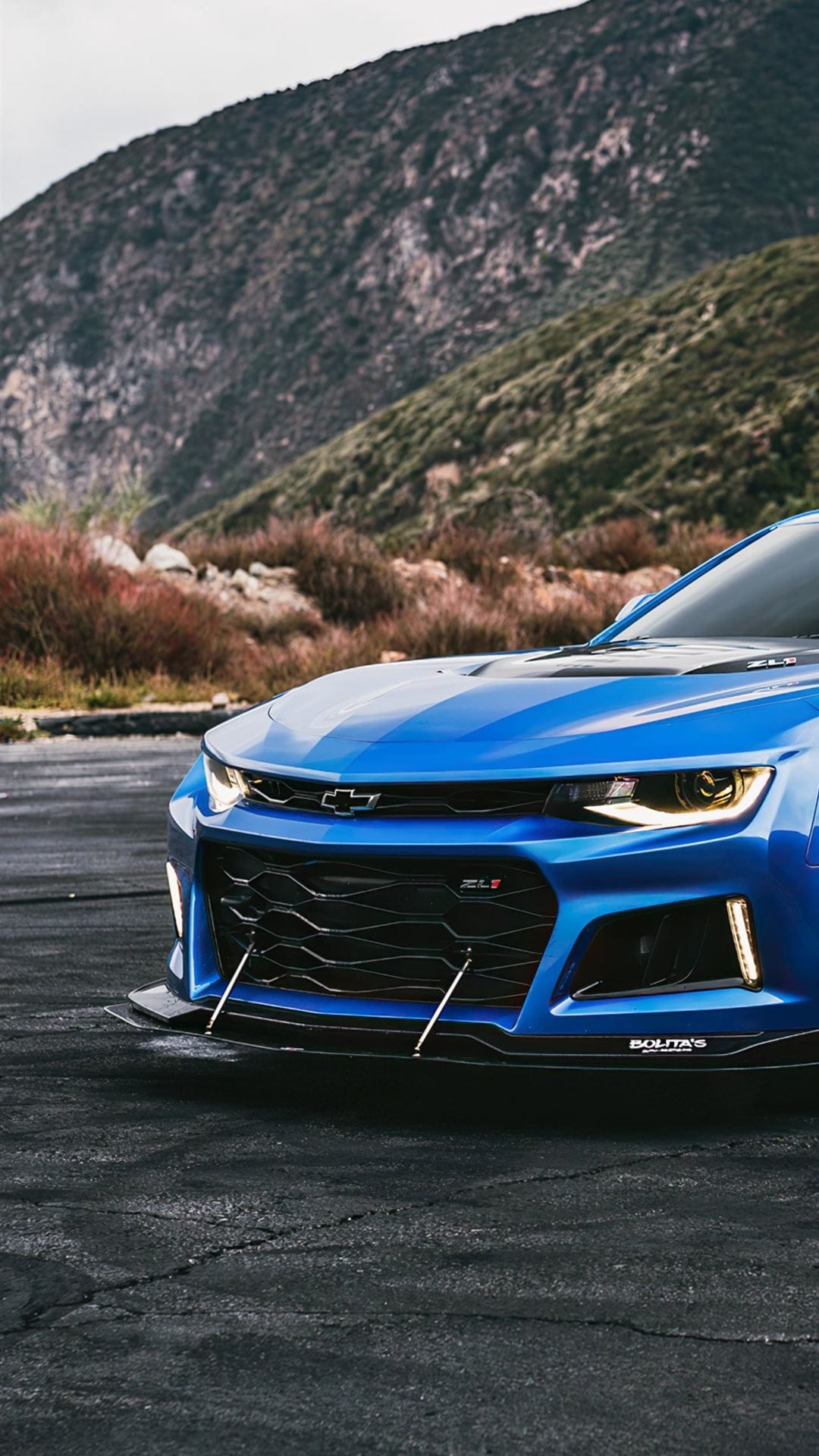 Blue Chevrolet Camaro ZL1 with lit headlights and a front splitter parked against a mountain landscape.