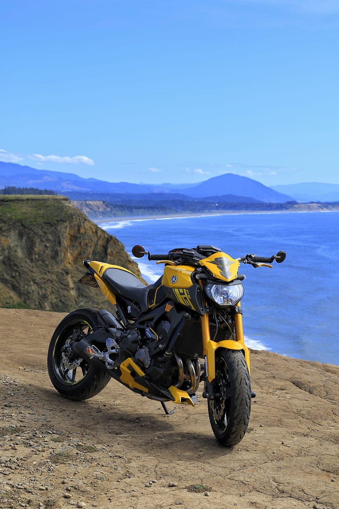 Yellow Yamaha motorcycle parked on a rocky cliff edge with a blue ocean and distant mountains in the background.