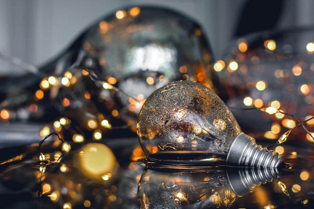 Glass Edison bulb ornament on a reflective surface surrounded by glowing golden fairy lights and blurred bokeh.