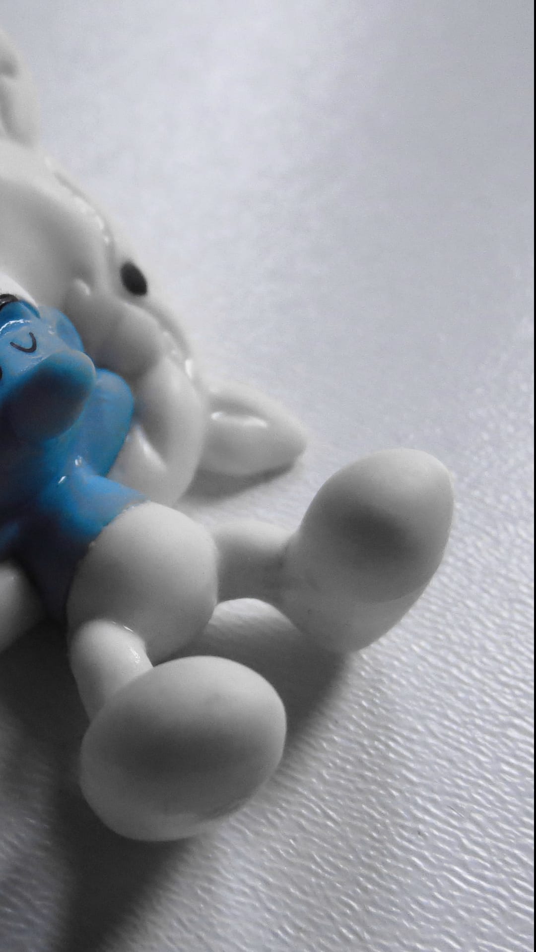 Blue Smurf toy figurine lies on its back on a white textured surface with a white toy rabbit in the background.