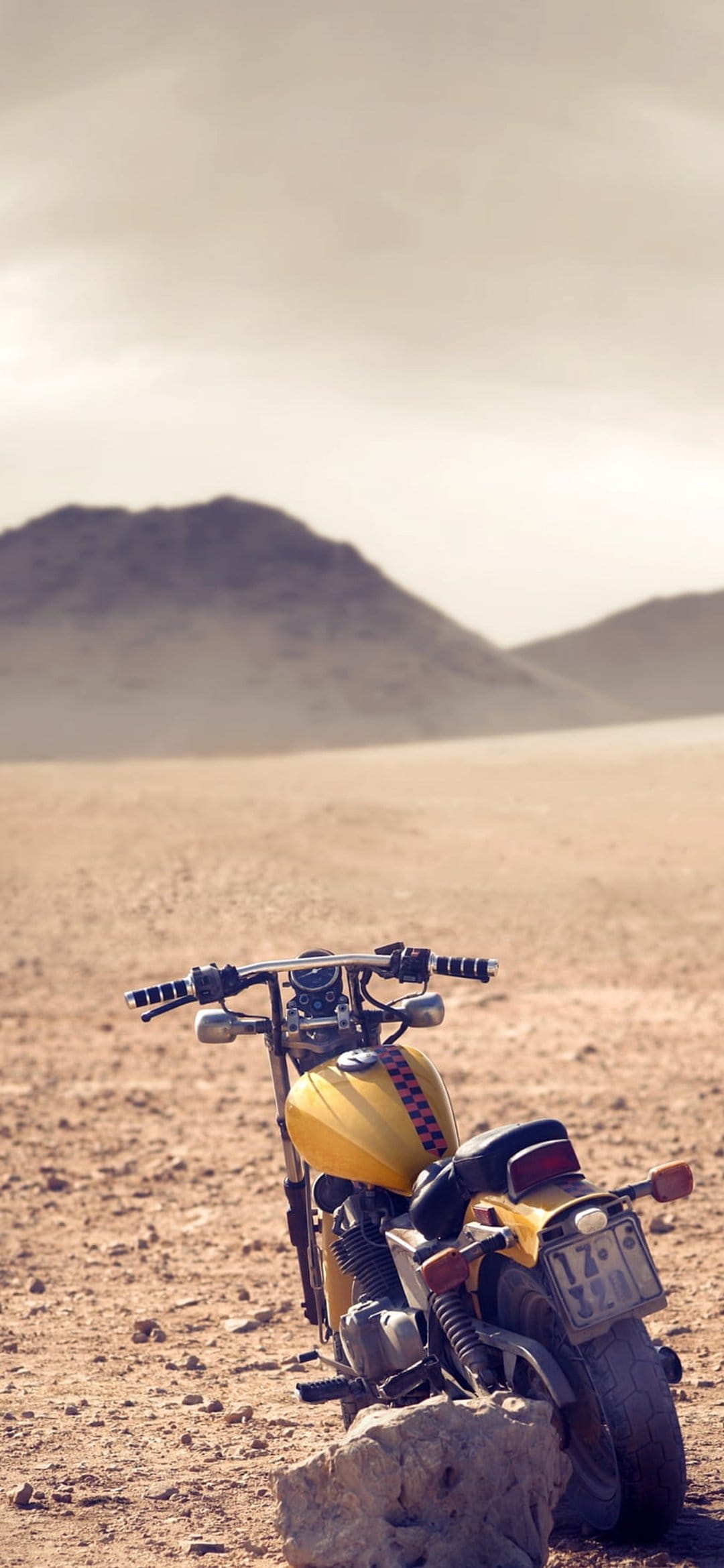 Yellow vintage motorcycle with a checkered fuel tank parked on sandy desert ground near hazy mountains.