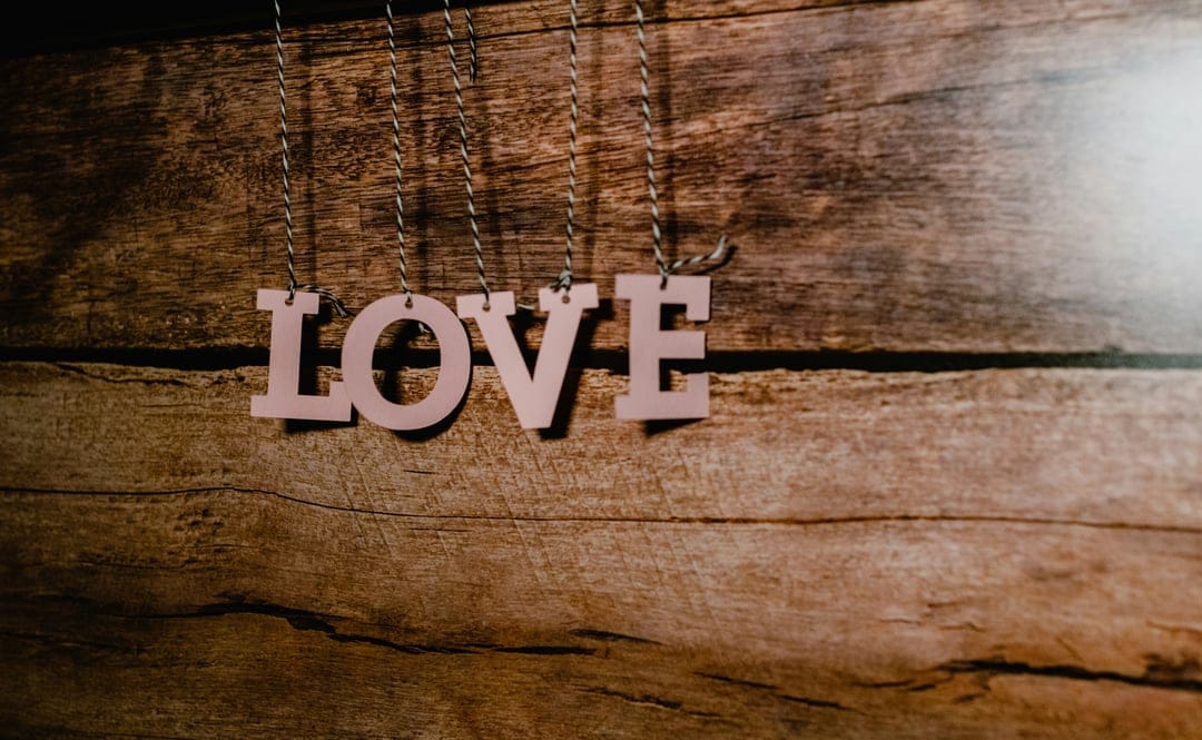 Pink wooden letters spelling love hang by strings against a textured brown rustic wood plank background.