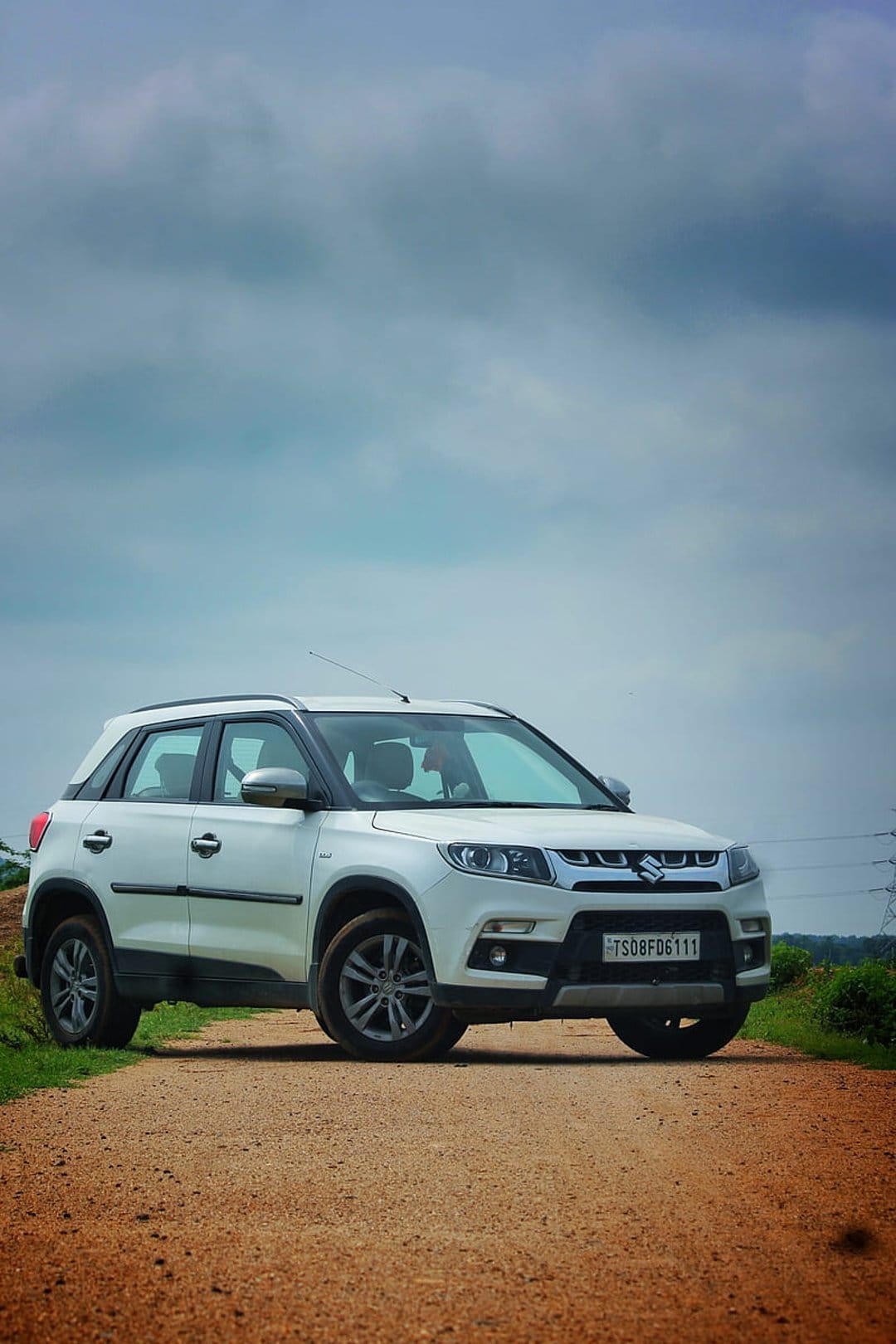 White Vitara Brezza SUV parked on a dirt road with alloy wheels visible under a dramatic overcast sky.