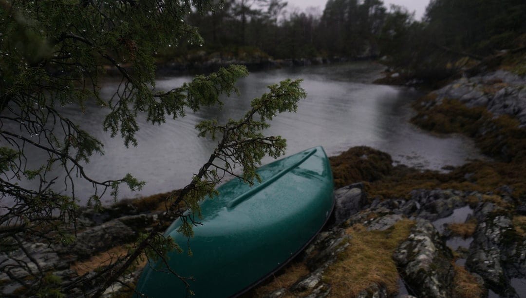 Green canoe rests on a mossy rocky shore beside a dark lake while rain falls in a misty, overcast pine forest.