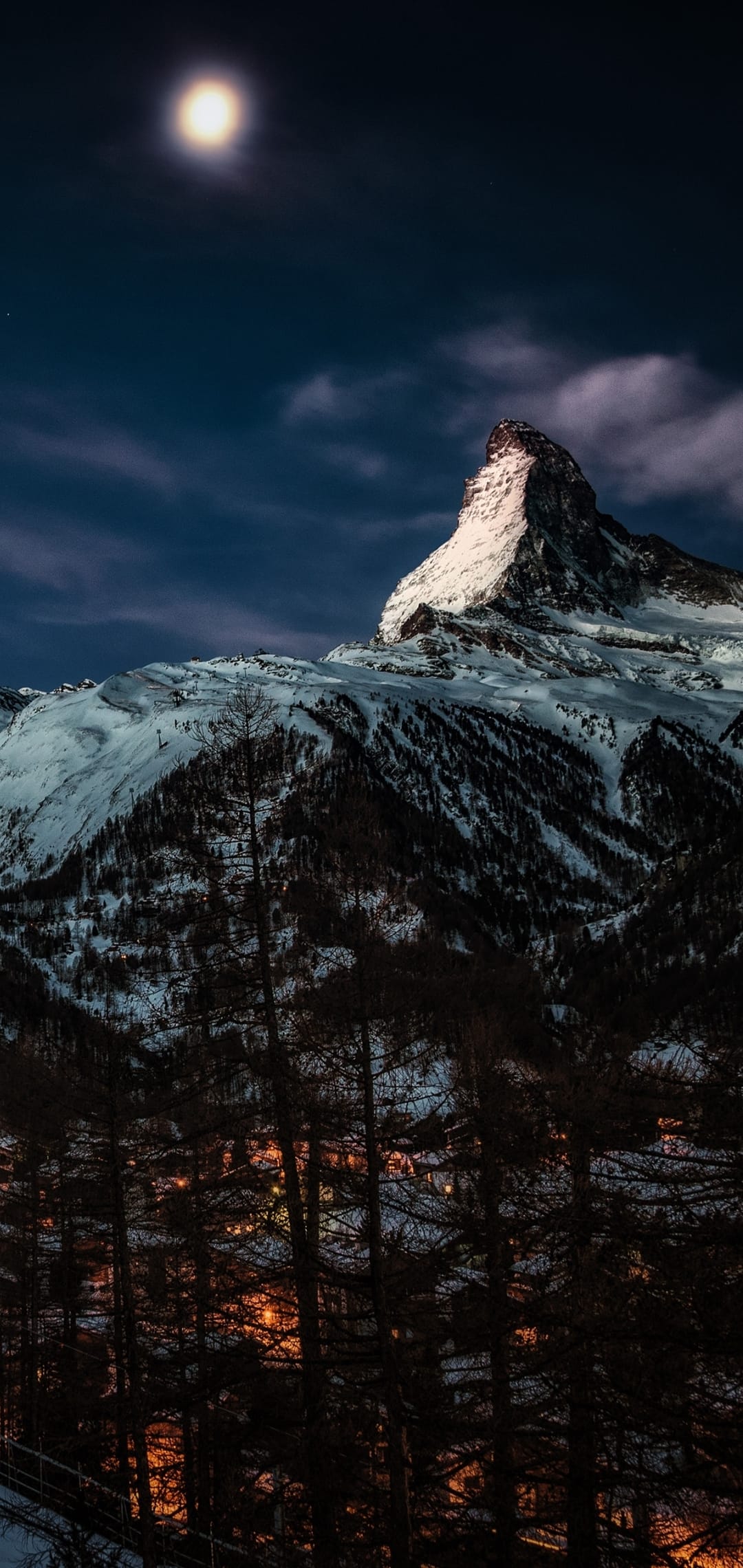 Matterhorn mountain peak glows under a full moon above the twinkling lights of an alpine village at night.
