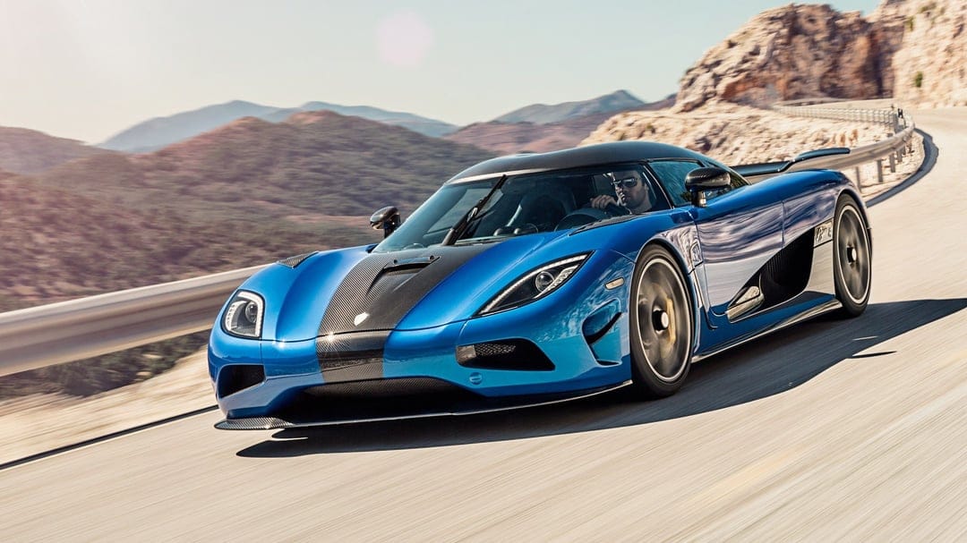 Blue Koenigsegg supercar driving fast on a winding mountain road with rugged hills in the background.