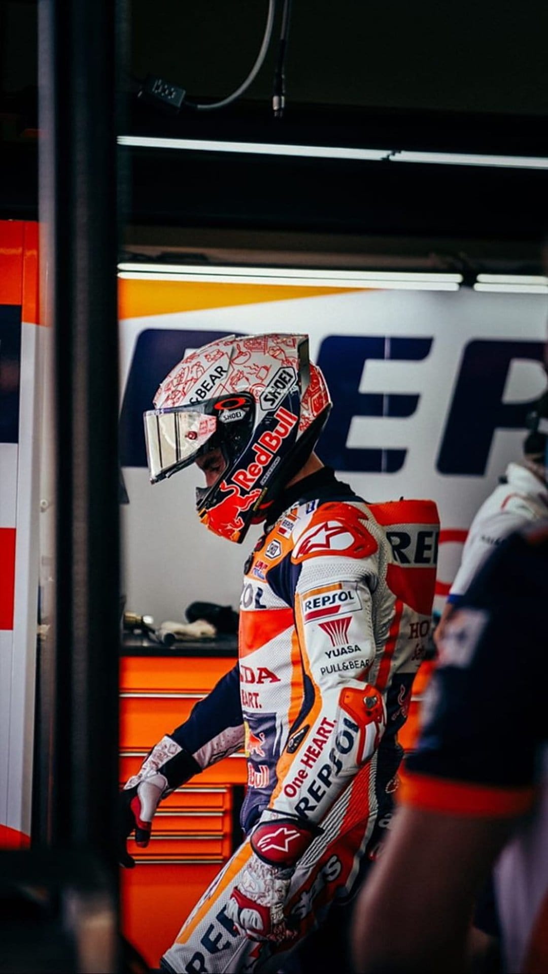 Marc Marquez wears a branded racing helmet and leather Repsol Honda suit while standing inside a dark pit garage.