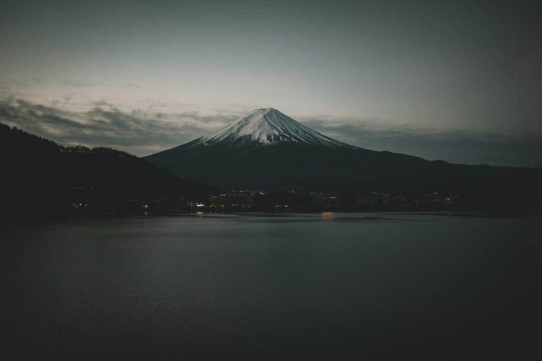 Mount Fuji with a snow-covered peak towers over a reflective lake during the purple and blue light of twilight.