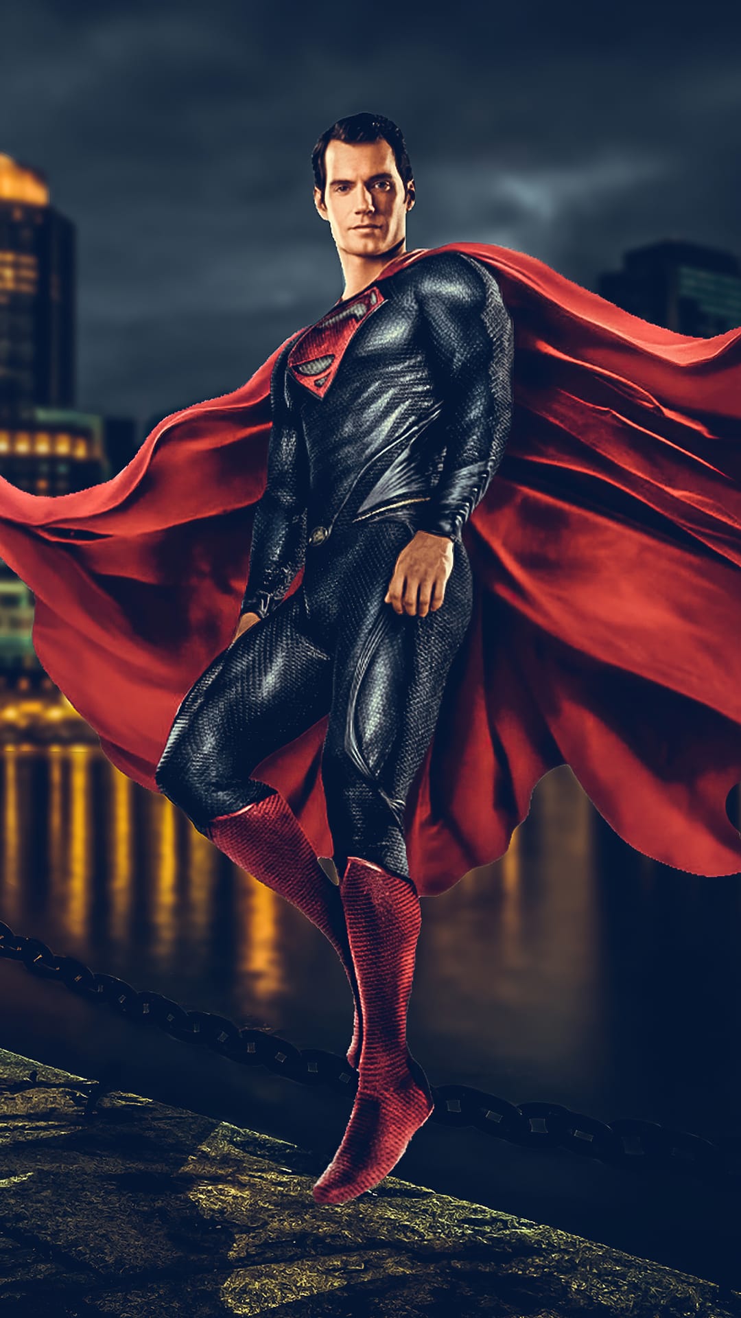 Superman stands on a rocky cliff overlooking a dark city with a long red cape billowing in the night wind.