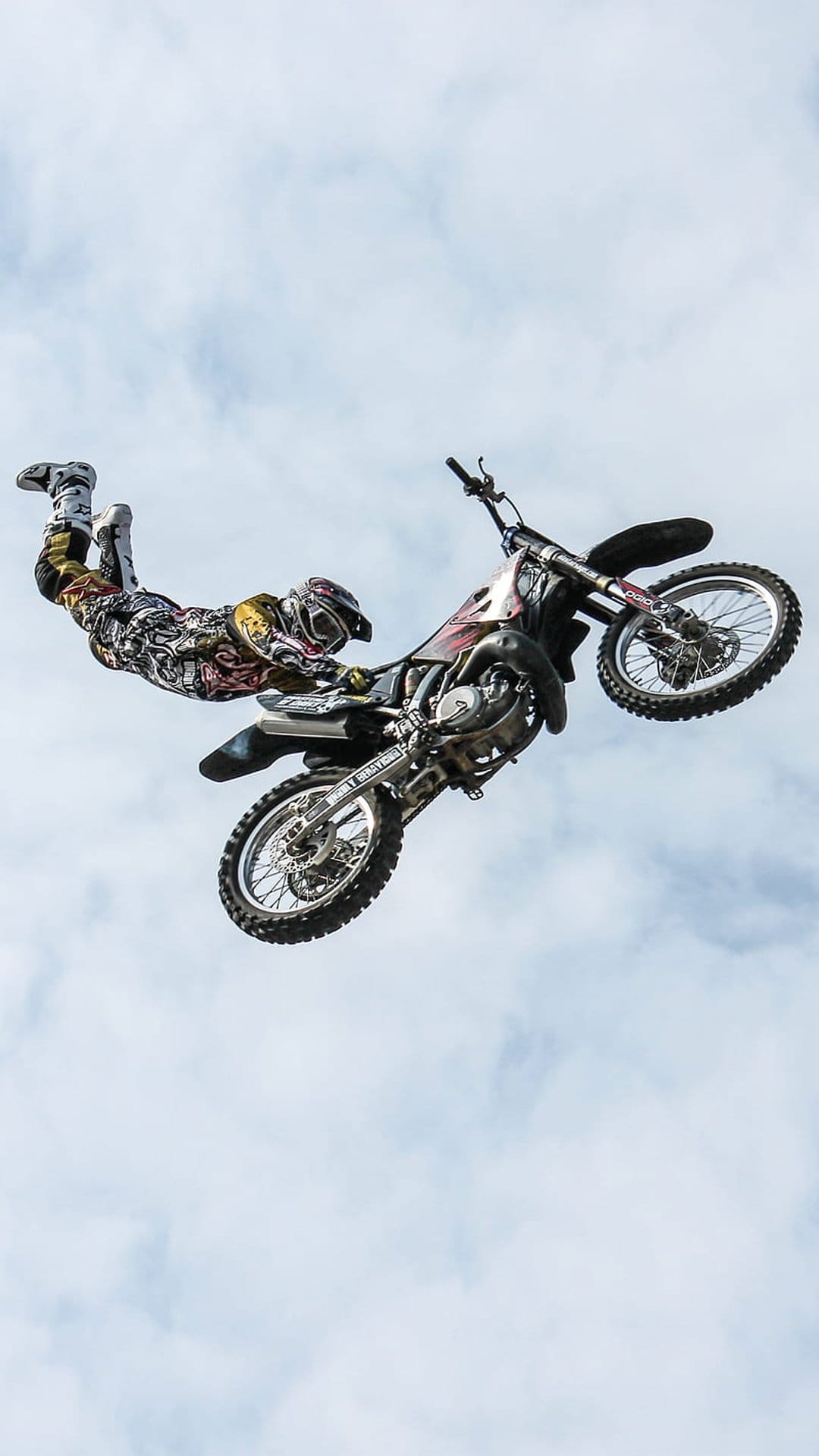 Motocross rider in mid-air performing a stunt on a dirt bike against a backdrop of soft white clouds in the sky.