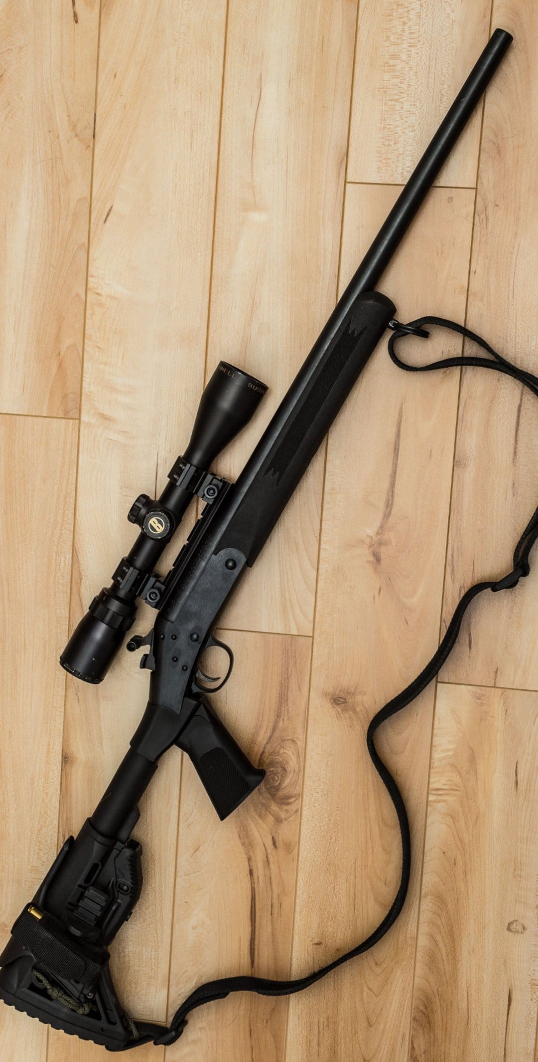 Black tactical rifle with a scope and sling lying horizontally on a polished light hardwood floor.
