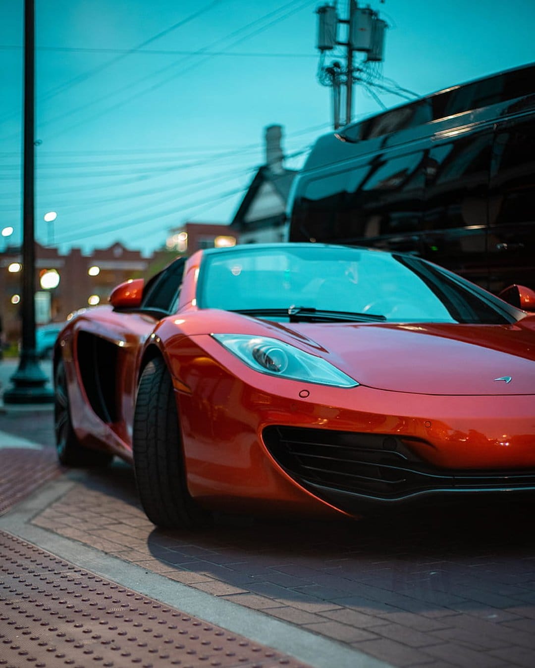 Red McLaren sports car with glowing headlights parked on a brick street against a teal twilight city sky.