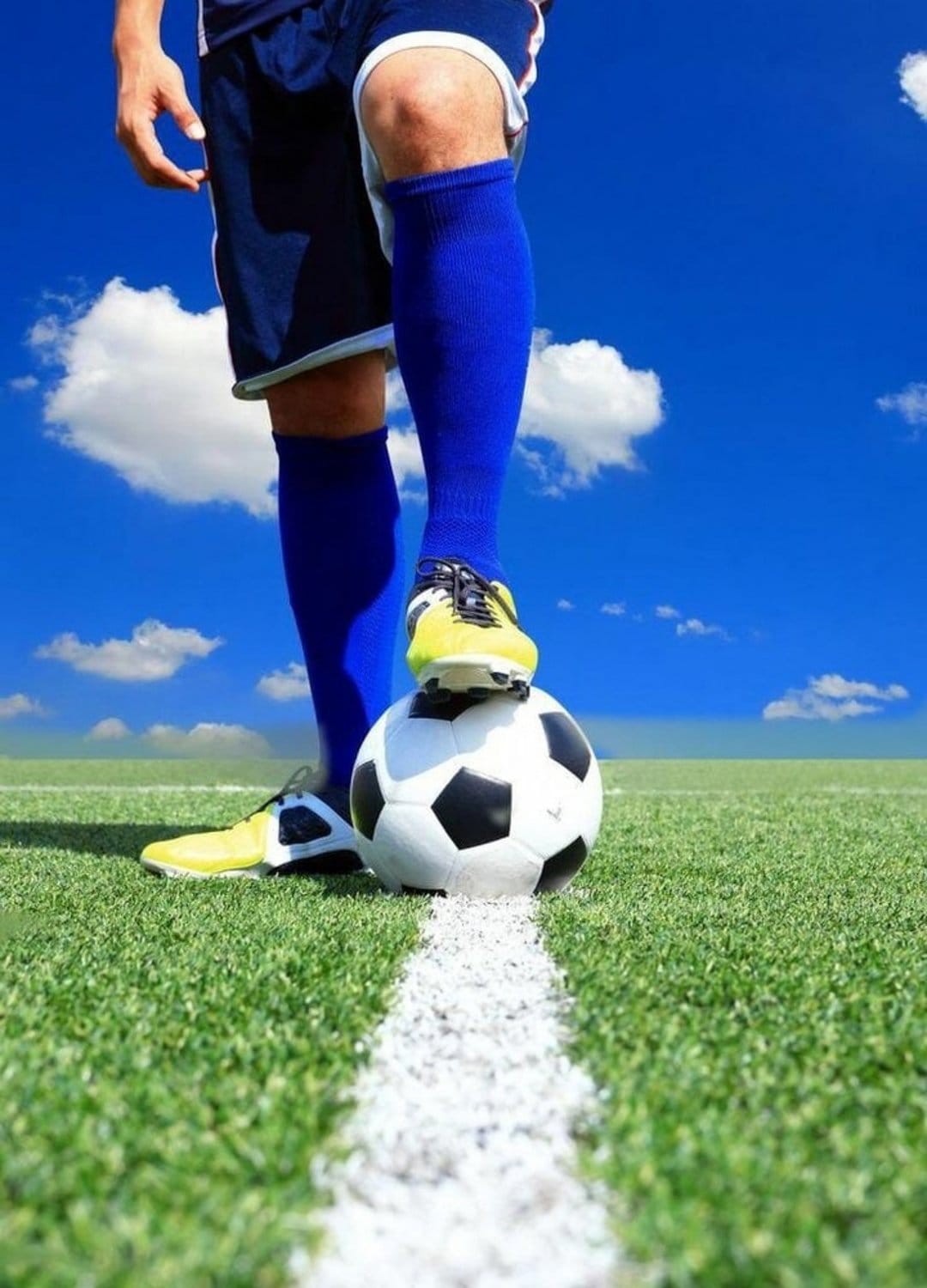 Soccer player wearing a yellow cleat and blue sock rests a foot on a football over a green grass pitch and sky.
