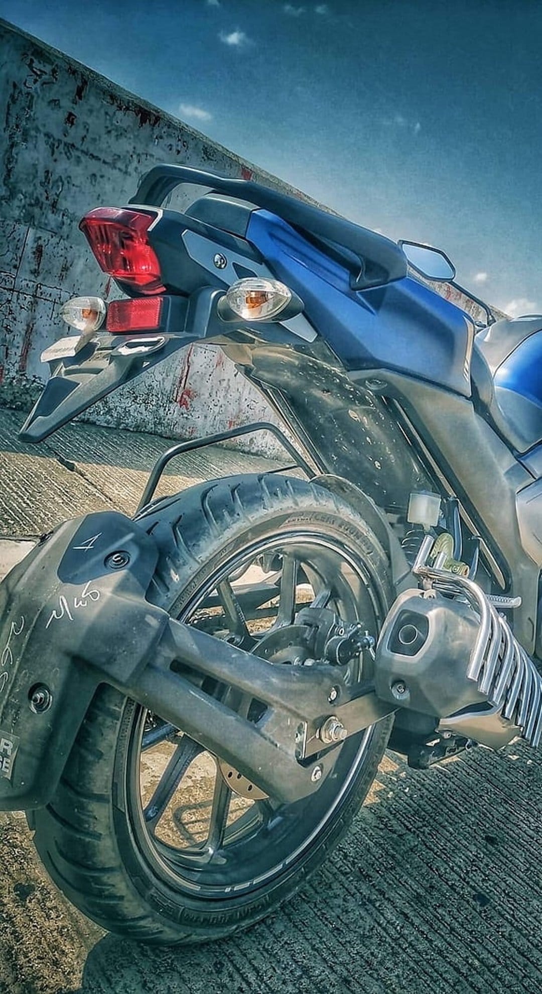 Blue motorcycle rear wheel with textured tire and red tail light against a concrete wall and cloudy blue sky.