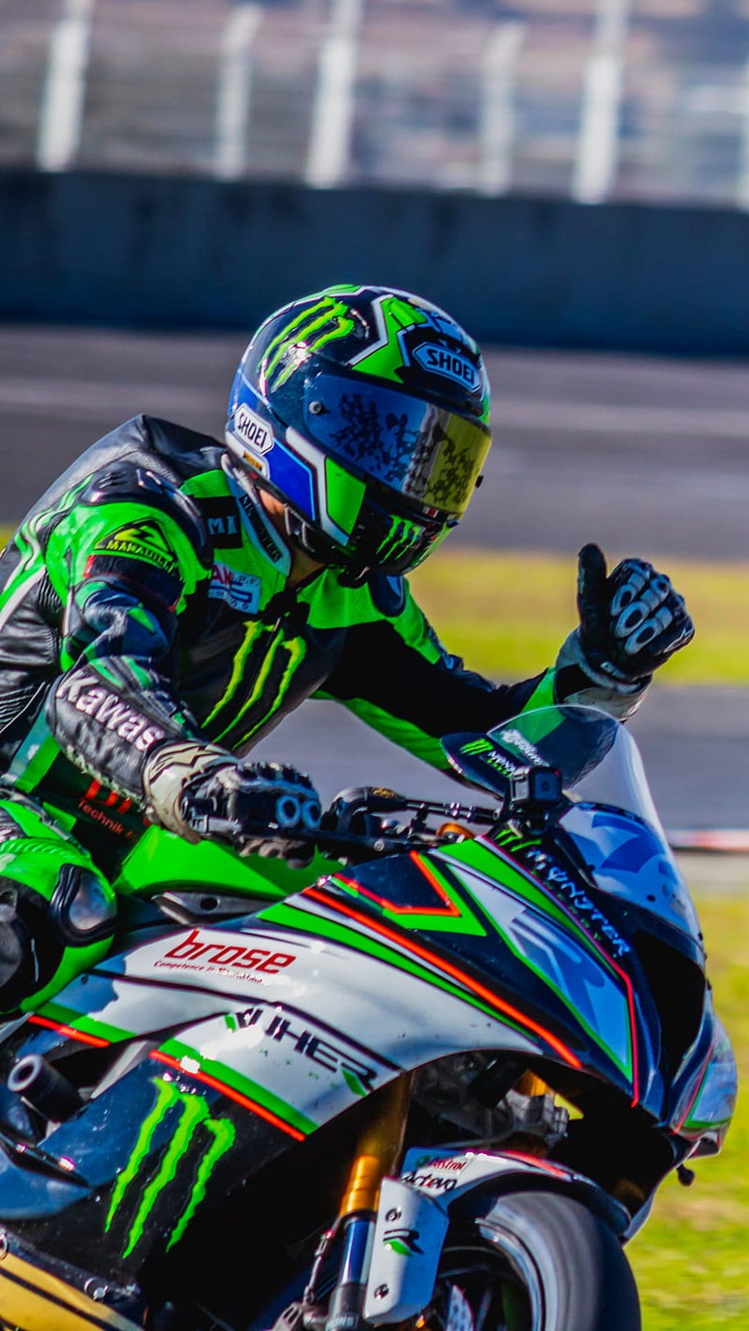 Professional racer in a green leather suit gives a thumbs up gesture wearing a black full-face helmet.