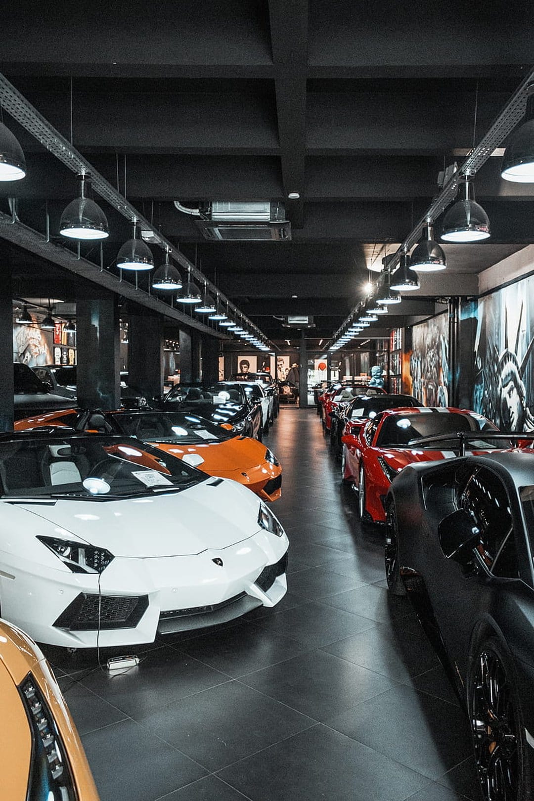 White and orange sports cars parked in a luxury showroom under industrial lights on a reflective floor.