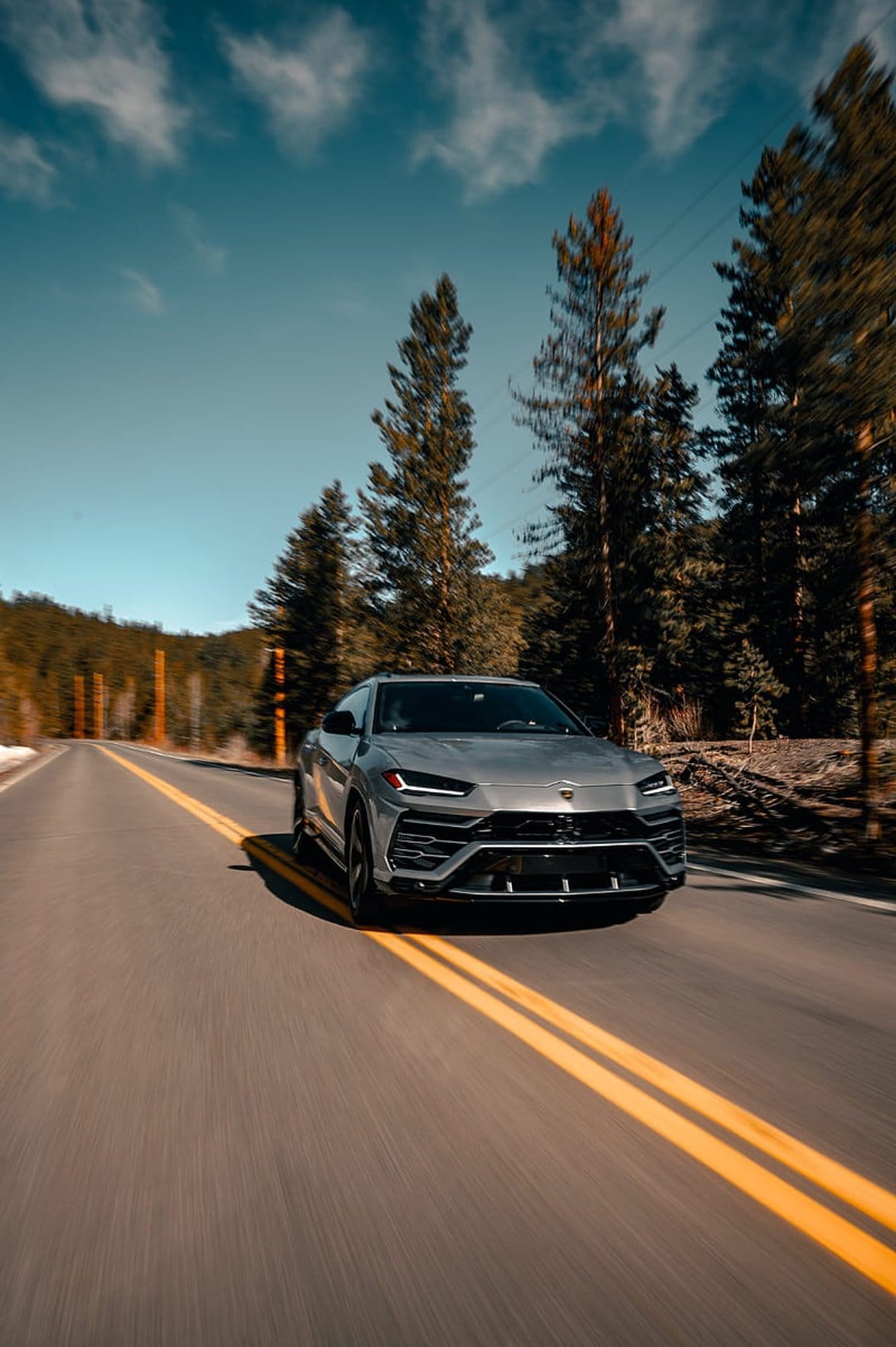 Silver Lamborghini Urus SUV speeds past evergreen trees on a mountain road with motion blur on the pavement.