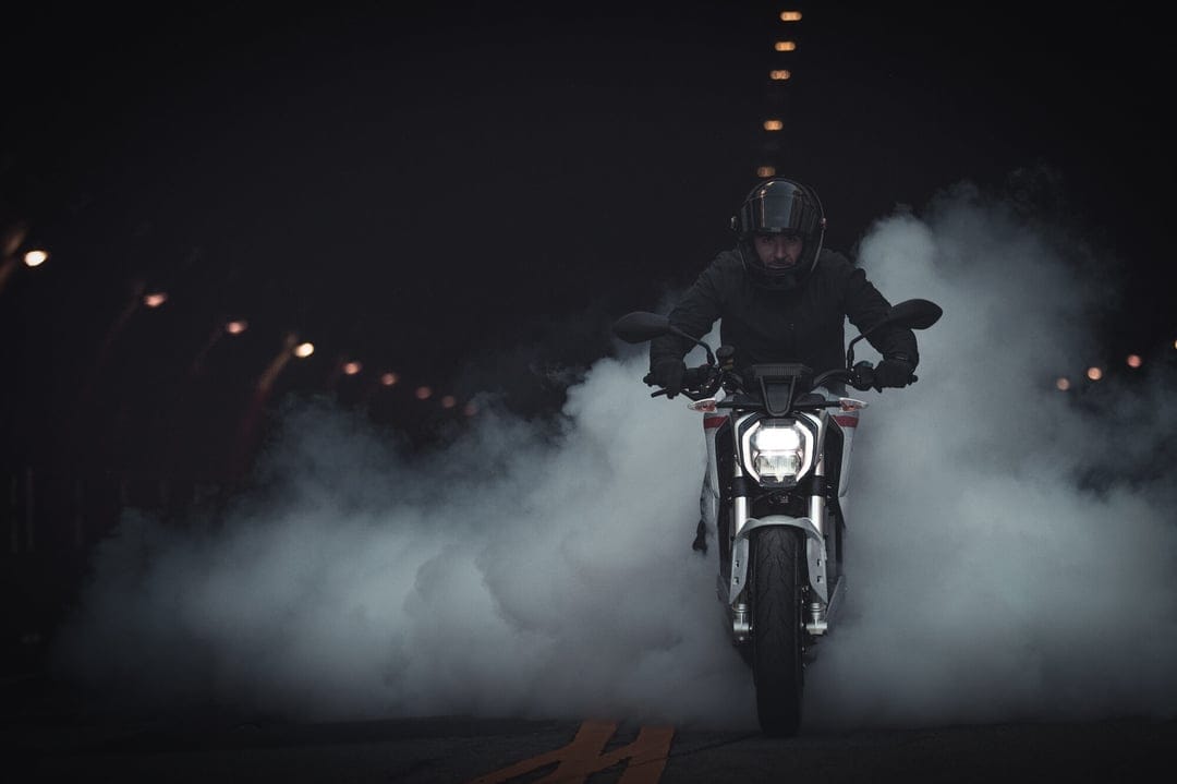 Motorcyclist in black gear performs a smoky burnout on a dark road with glowing headlights and bokeh city lights.