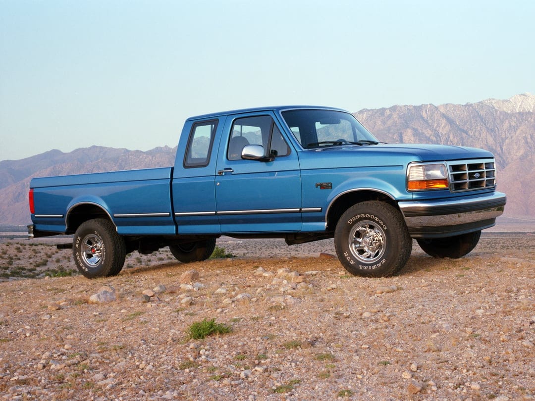 Blue Ford F-150 SuperCab parked on rocky desert ground with towering mountains and a clear sky in the background.