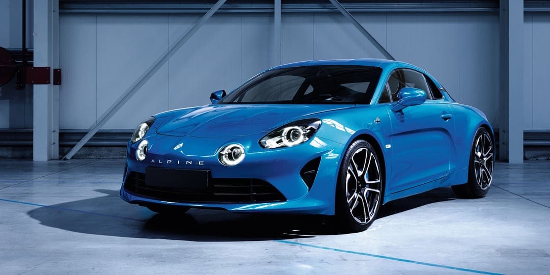 Blue Alpine A110 sports car with glowing LED headlights parked on a concrete floor in a gray industrial studio.