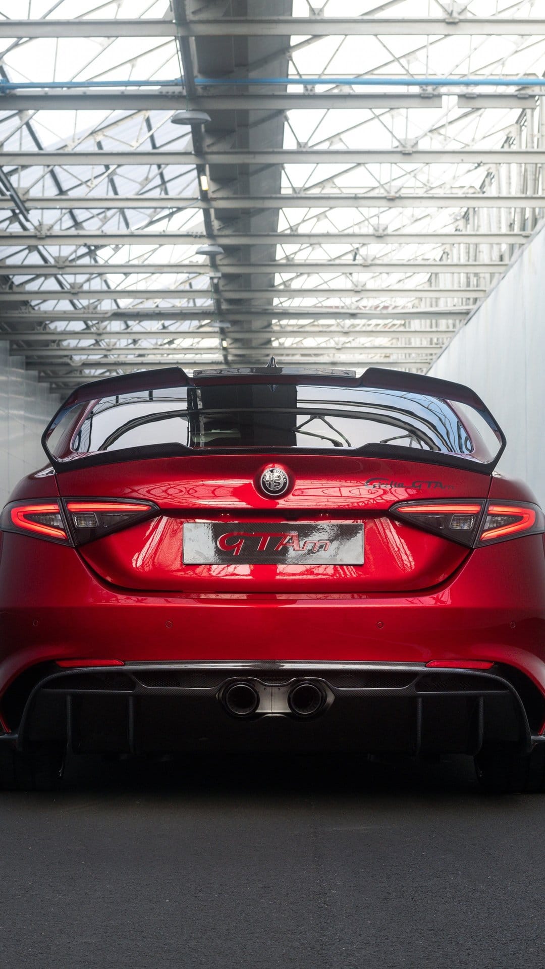 Red Alfa Romeo Giulia GTAm rear with a large carbon wing, lit taillights, and dual exhausts in a metal warehouse.