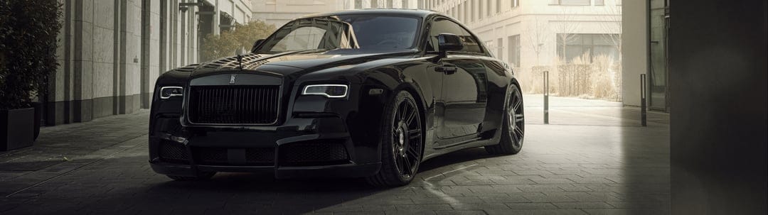 Front view of a black Rolls-Royce Wraith coupe parked in a sunlit urban alley with modern stone buildings.