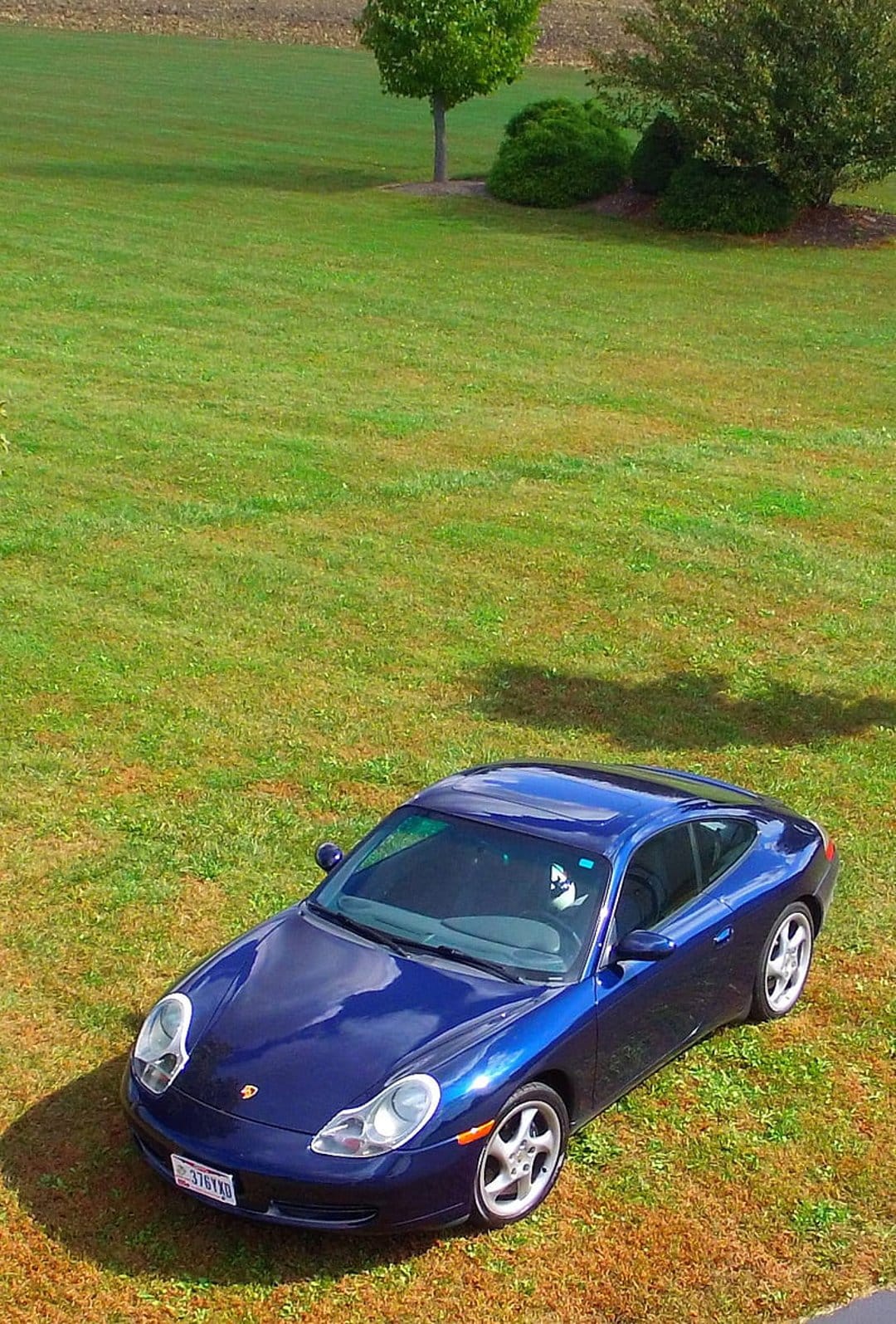 Dark blue Porsche 911 coupe parked on a green grass lawn with reflections of the sky and trees on its paint.