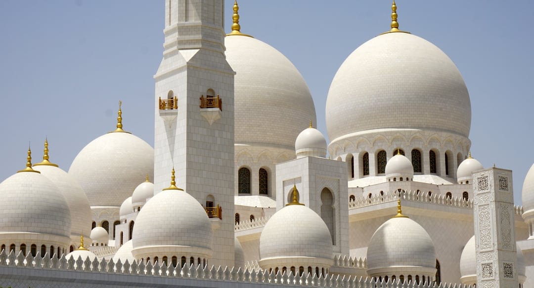 White marble domes and a tall minaret of the Grand Mosque rise into a clear blue sky during bright daylight.