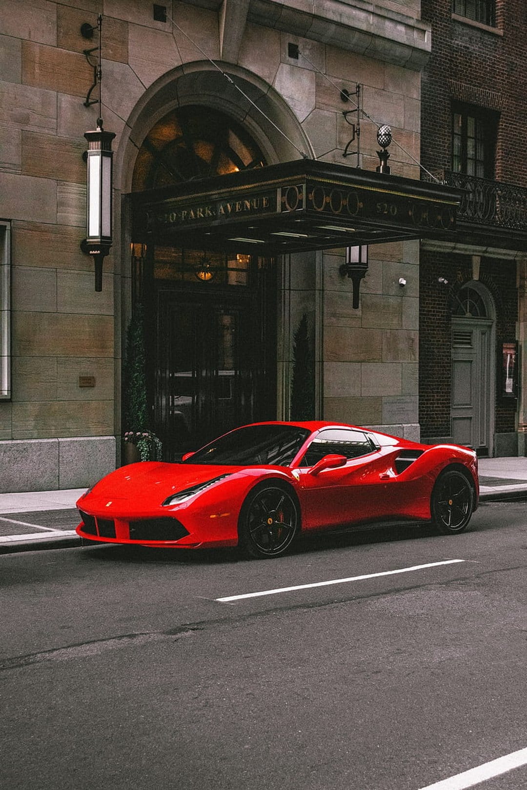 Red Ferrari sports car parked by the ornate stone entrance of a luxury Manhattan building in an urban setting.