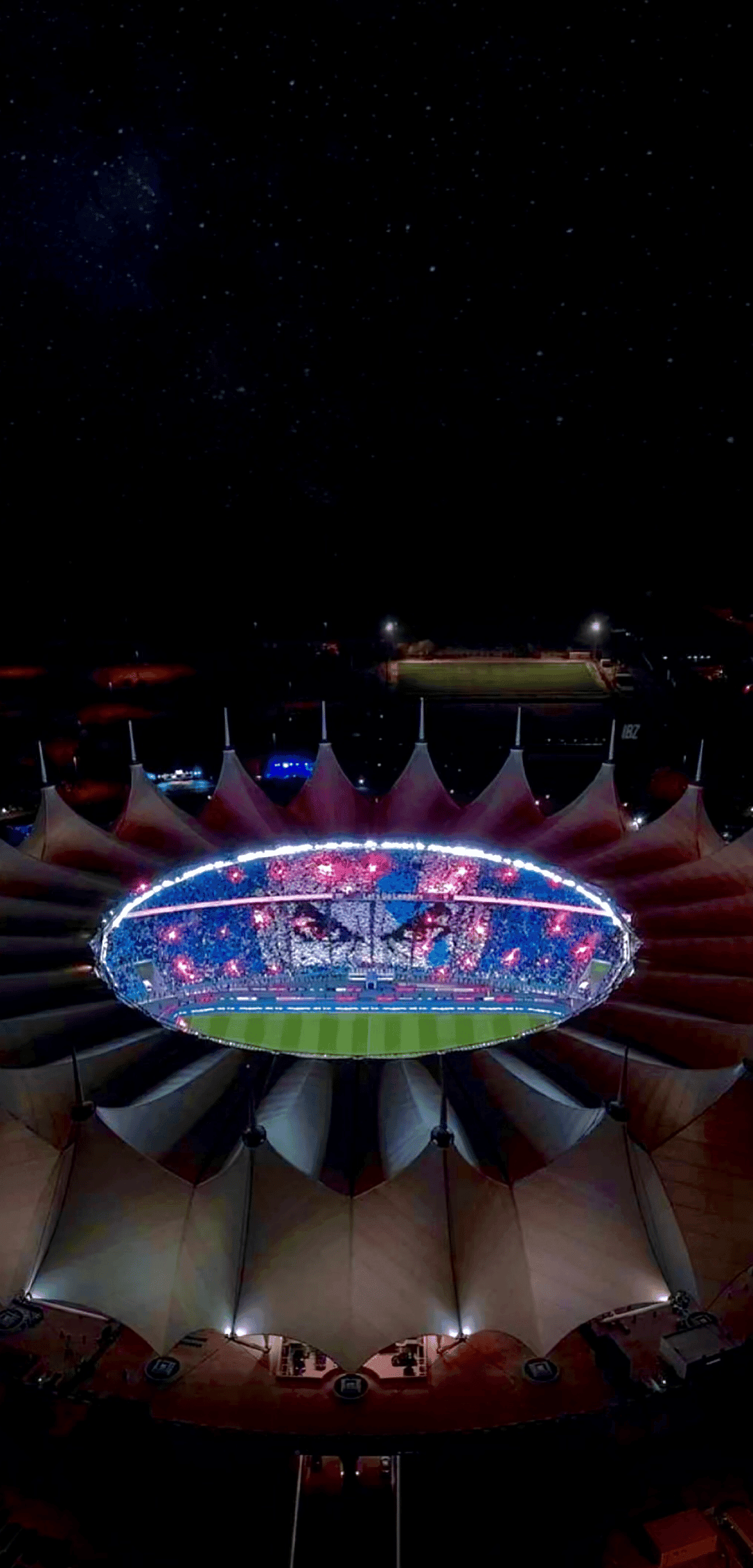 Football stadium with a glowing tent-roof and green field shines under a starry night sky from an aerial view.