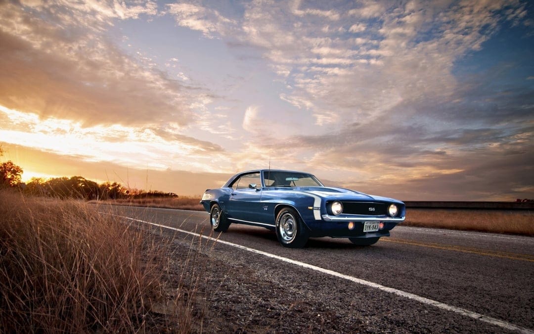 Blue 1969 Chevrolet Camaro SS muscle car parked on a road at sunset with dramatic clouds and golden lighting.