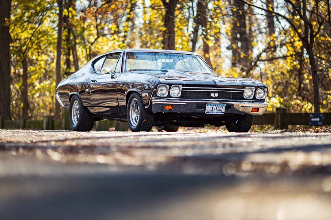 Black classic Chevelle SS 396 muscle car on a rural road surrounded by golden autumn trees in sunlight.
