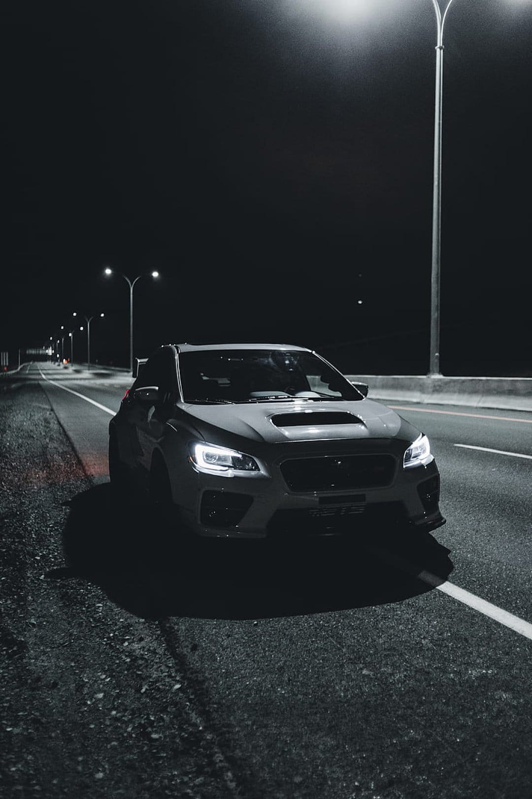 White Subaru WRX with glowing headlights parked on a dark highway under streetlights in a vertical composition.
