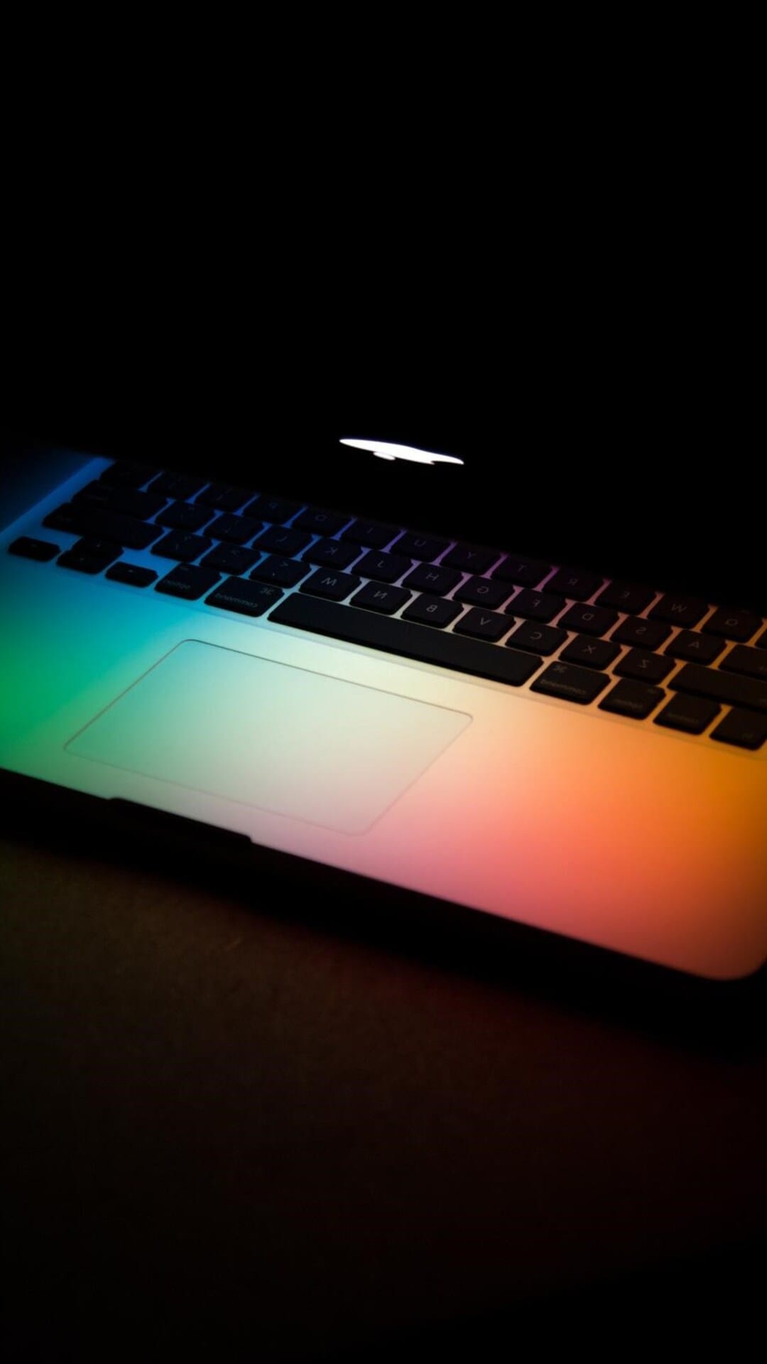 Laptop keyboard and trackpad glowing with a rainbow LED gradient from green to red on a dark surface.