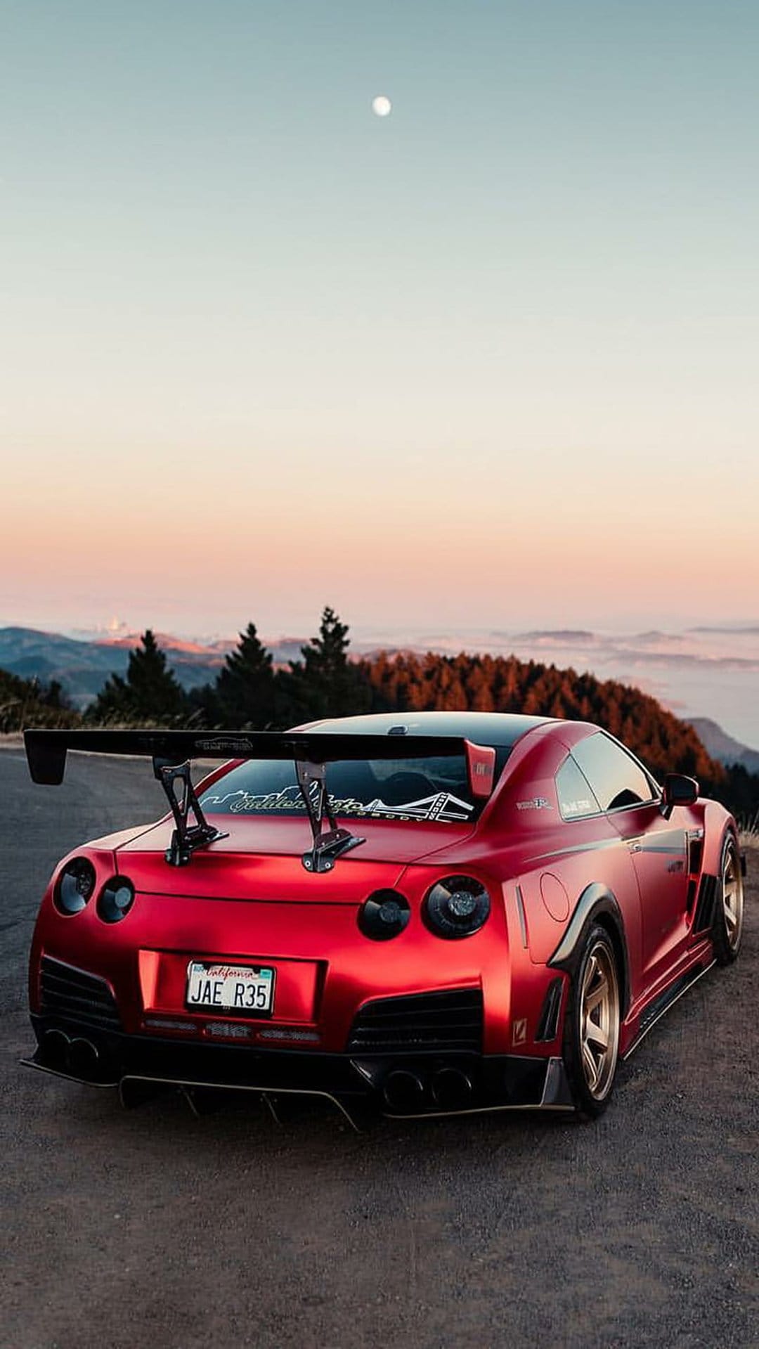 Red Nissan GT-R with a large rear wing parked on a gravel mountain path under a vibrant orange sunset sky.
