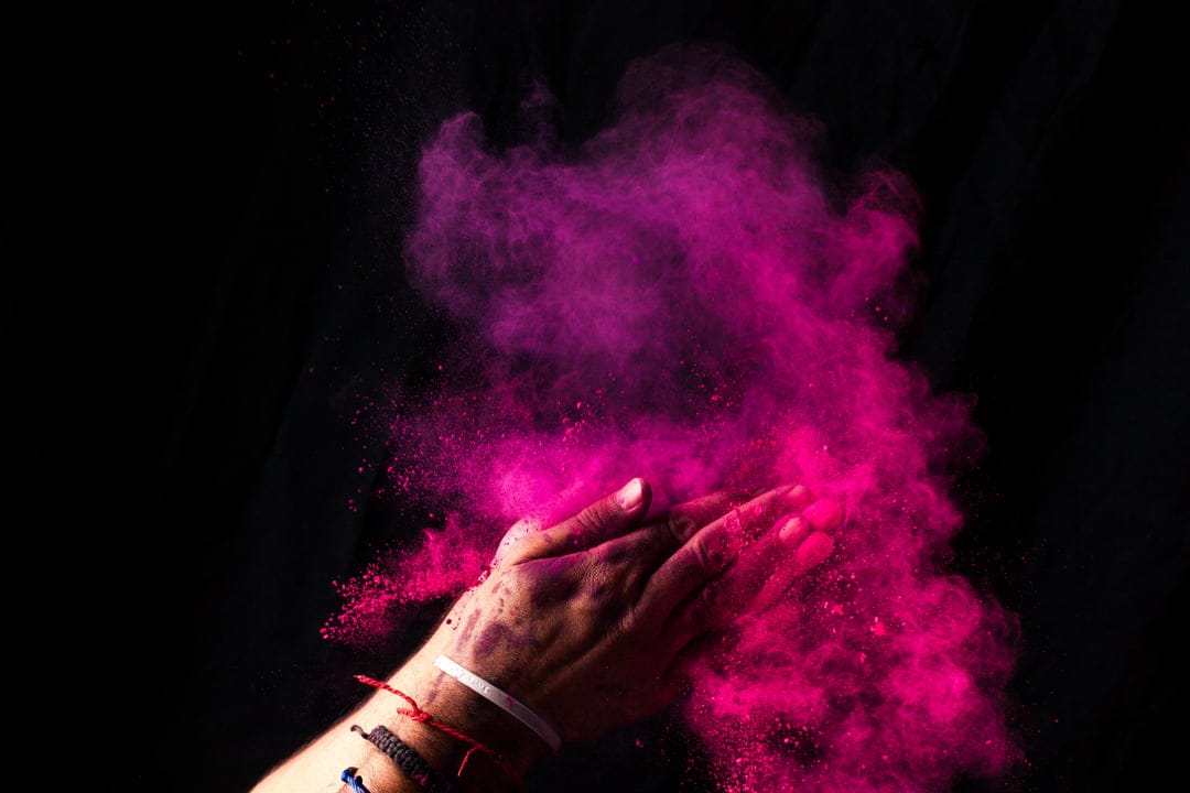 Two hands release a vibrant cloud of pink Holi powder with a smoky texture against a dark black background.