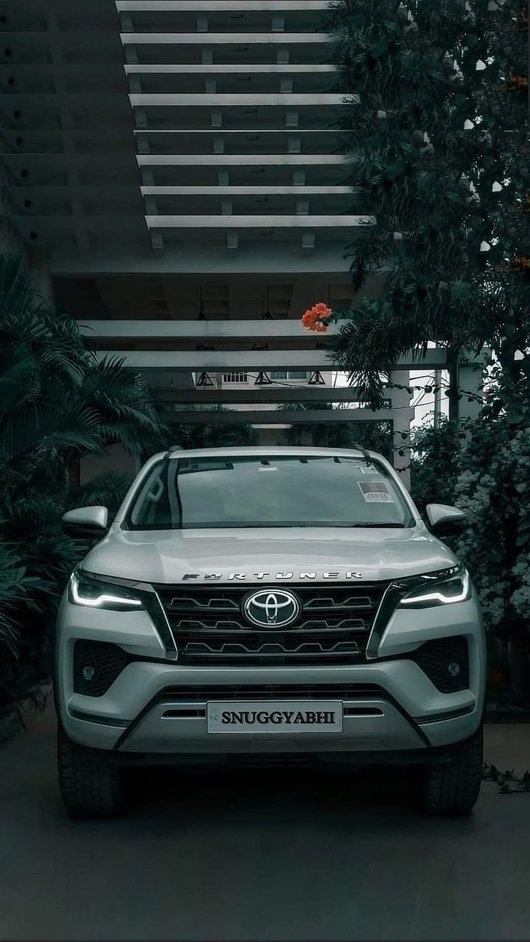 Toyota Fortuner SUV with glowing LED headlights parked near a modern building and lush green plants.