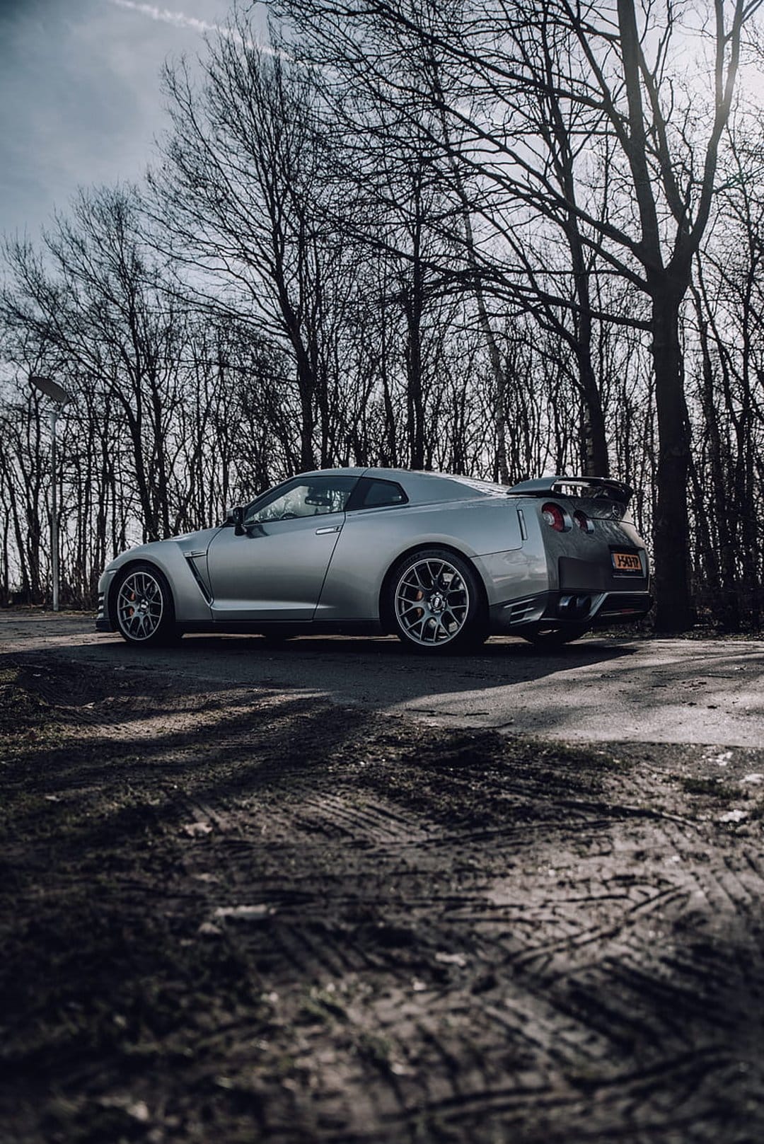 Silver Nissan GT-R sports car parked on a dirt road between bare trees under a cloudy, atmospheric gray sky.