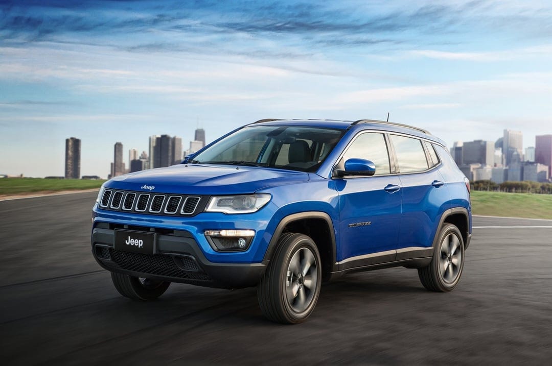 Blue Jeep Compass SUV driving along a paved road with a distant city skyline under a bright, cloud-streaked sky.